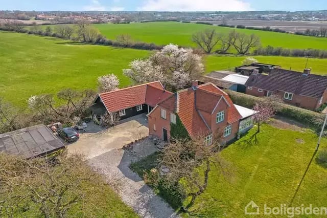 A red brick house on a courtyard surrounded by green fields and blossoming trees. Cars are parked next to the house, which is located in a rural area.