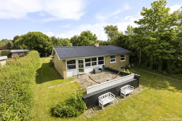 A yellow brick house with a black roof, set in a green garden surrounded by bushes. In front is a terrace with two white benches by a black fence. Sunny weather.