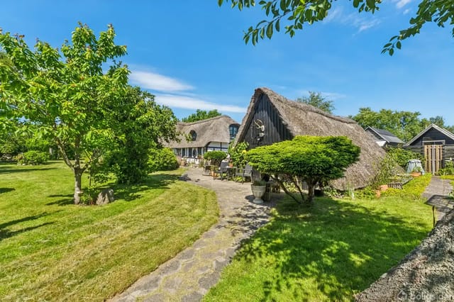 Thatched house surrounded by lush garden with lawn and trees; a stone path leads to a terrace with garden furniture. Sunlight creates an idyllic atmosphere.