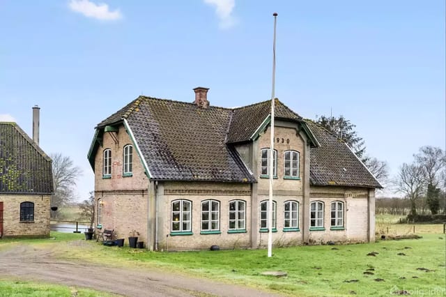 A charming old brick house with a sloped roof, built in 1930, standing on a lawn with a flagpole in front, surrounded by open landscape and a smaller building in the background.