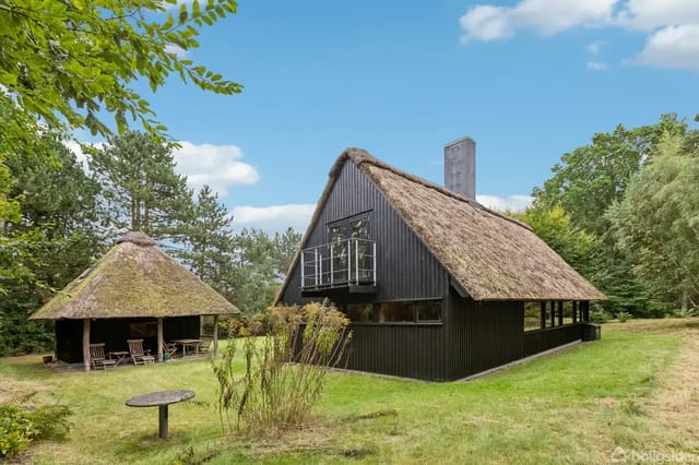 Thatched house in a lush garden with surrounding trees and a visible terrace.
