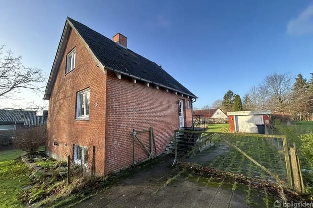 Brick house with pitched roof in a sunny garden. Concrete steps lead to the entrance. Surrounding lawn and bushes encircle the property, with sparse trees in the background.