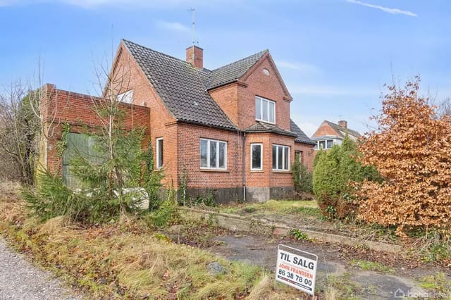 A red brick villa for sale, surrounded by a garden with trees and bushes. In front of the house is a sign with the text: 'FOR SALE 86 38 78 00 real estate agency'.