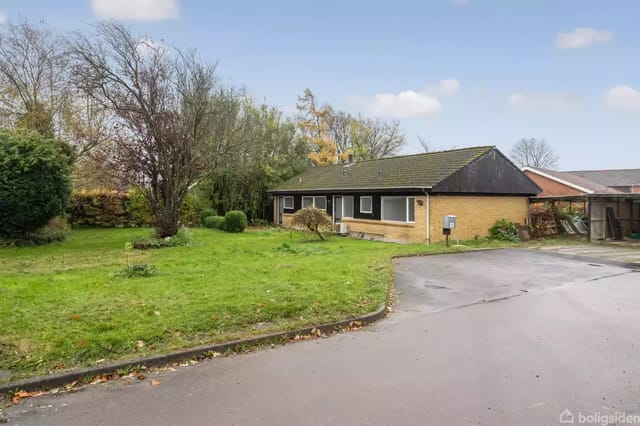 Yellow brick bungalow surrounded by a green garden with trees and bushes.