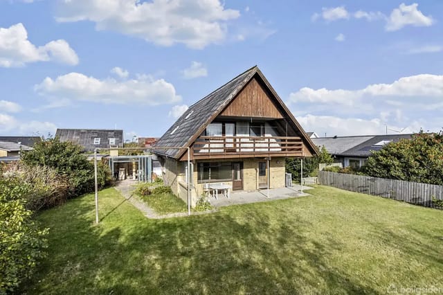 A-shaped house situated in a green garden with lawn and trees in a residential neighborhood. The house features a balcony and large windows in the roof.