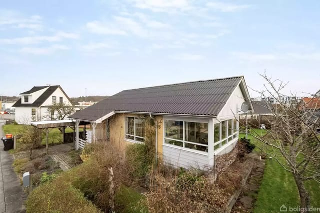 A simple house with a gray roof and large windows surrounded by a well-kept garden. Neighbors can be seen in the background under a clear blue sky.