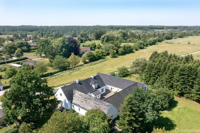 A large white house with a dark roof surrounded by lush green vegetation, set in a rural area with forests and fields in the background.