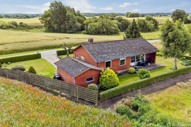 Red brick house with flat roof in a rural garden surrounded by fields and bushes, with a road in the background under a clear sky.