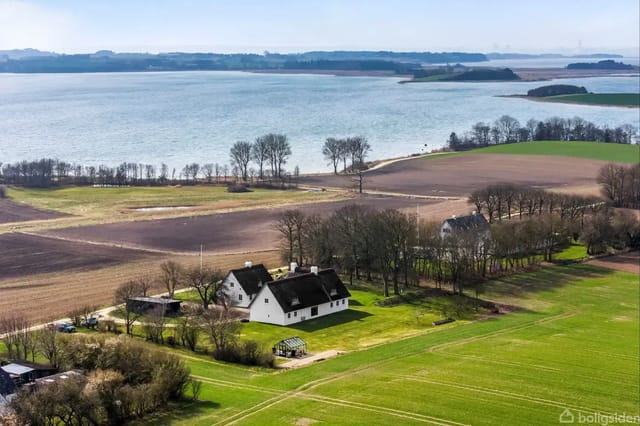 A white country house with thatched roof among trees and fields, overlooking a bay.