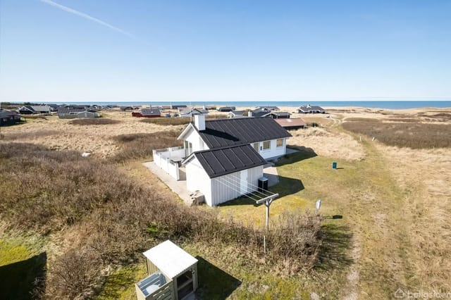 A white holiday home with a black roof surrounded by grass and dunes, with several houses in the background and the sea stretching along the horizon.
