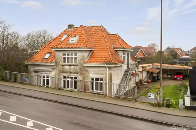 A large house with a red tile roof stands by a road. The house has an external staircase and several windows. It is surrounded by a garden and neighboring houses in a residential area.