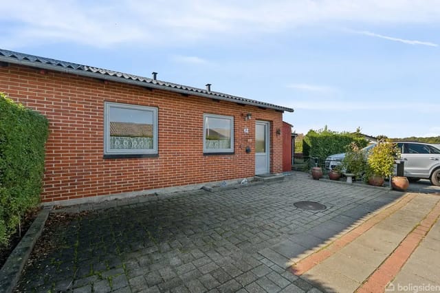 A red brick building with two windows and a door stands on a paved courtyard, surrounded by a trimmed hedge and potted plants. A car is partially visible to the right.