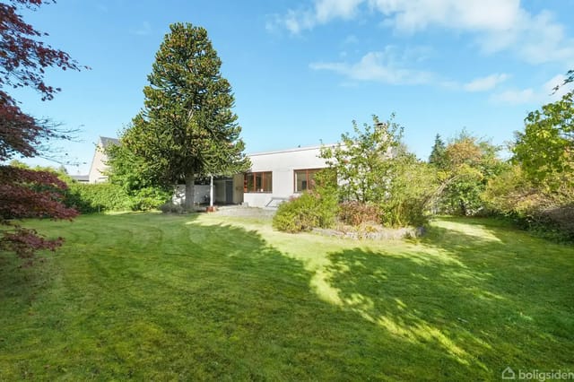 A white house stands quietly in a large, green garden surrounded by trees and bushes. Sunlight shines on the lawn, and the sky is clear and blue.