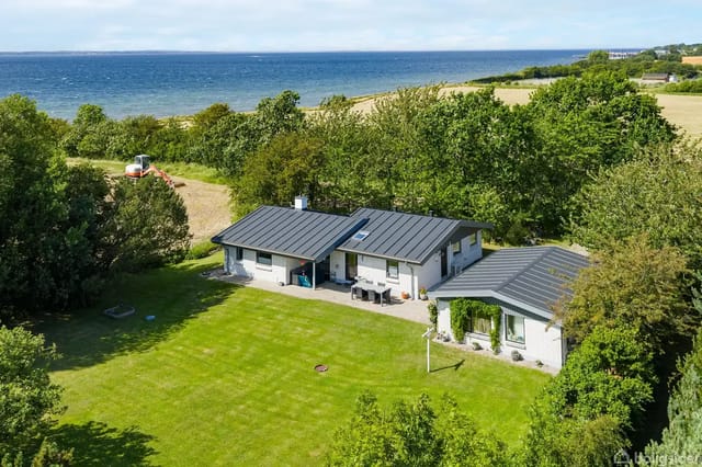 The house sits quietly in a green garden with a large lawn, surrounded by trees. In the background, the sea is visible under a blue sky.