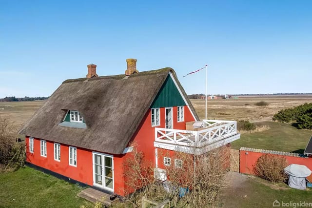 A red thatched house with two chimneys stands in an open landscape. A large terrace with railing is visible on the side. Blue sky in the background.