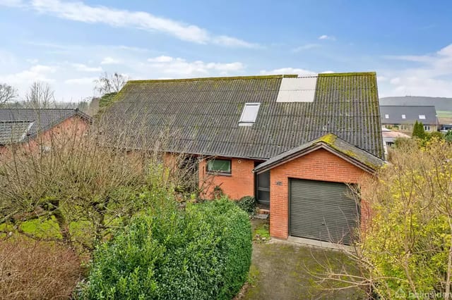 Brick house with sloped roof, garage door, and two windows, surrounded by trees and bushes in a residential area. Clear sky with few clouds.