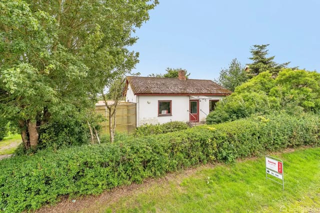 A small white house with a red roof surrounded by green trees and bushes. A 'For Sale' sign from the real estate agency Nybolig is visible in front of the house.