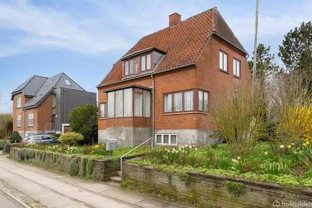 A red brick house with a sloping roof and several windows, surrounded by a lush garden with flowers. The house stands by a roadside with another house in the background.