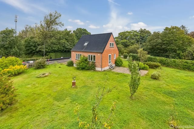 A red brick house with a black roof stands in a green garden with bushes and trees. A road is seen in the background, surrounded by dense forest.