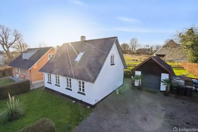 A white house with a red tile roof stands in the sun next to a garage on a gravel road. Around the house is a green garden and some trees in the background.