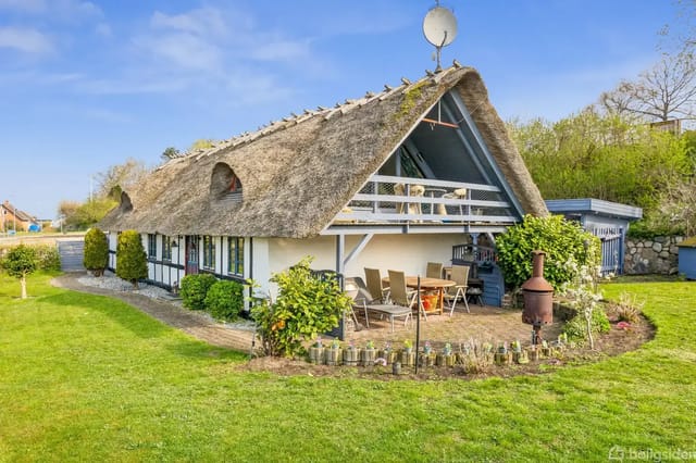 Thatched house with sun loungers on the terrace, surrounded by a well-kept garden. A satellite dish is mounted on the roof, and there are trees in the background.