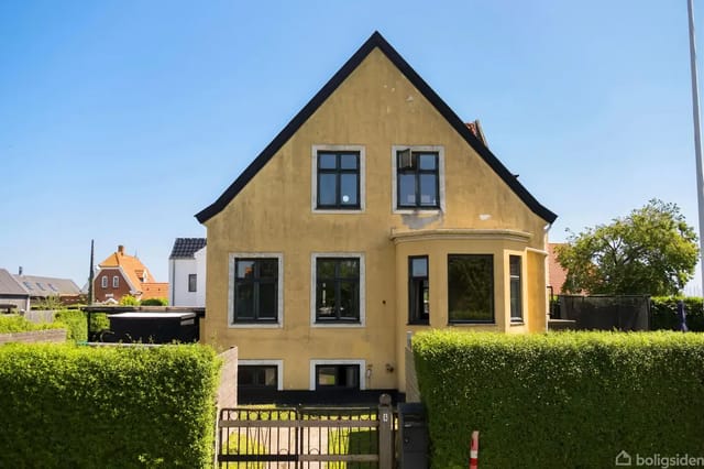 A yellow house with a black roof surrounded by a well-kept hedge in a residential area. The house has several large windows and a small front yard with an open gate.