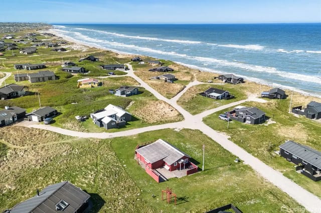 Holiday homes scattered along a coastline with beaches and waves, set on grassy areas connected by small roads, with the sea in the background.