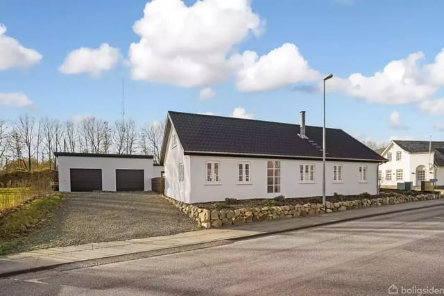 White house stands still by the country road, surrounded by a low wall and gravel driveway. In the background, a garage and trees under a blue sky with few clouds.