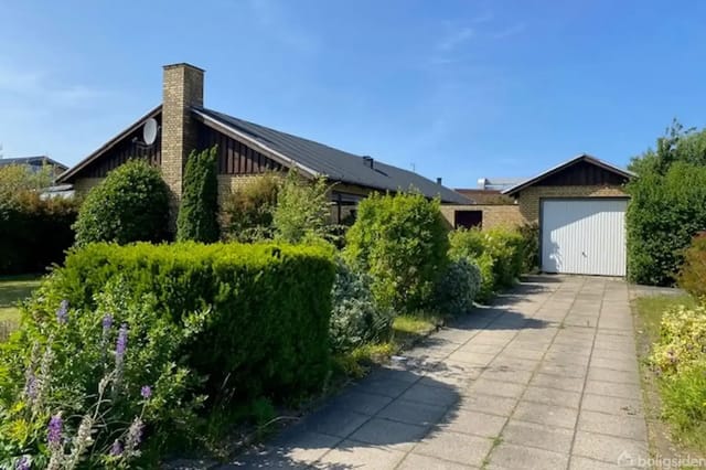 A single-family house in red brick surrounded by a well-kept garden with shrubs and flowers, next to a paved driveway and garage in sunny weather.