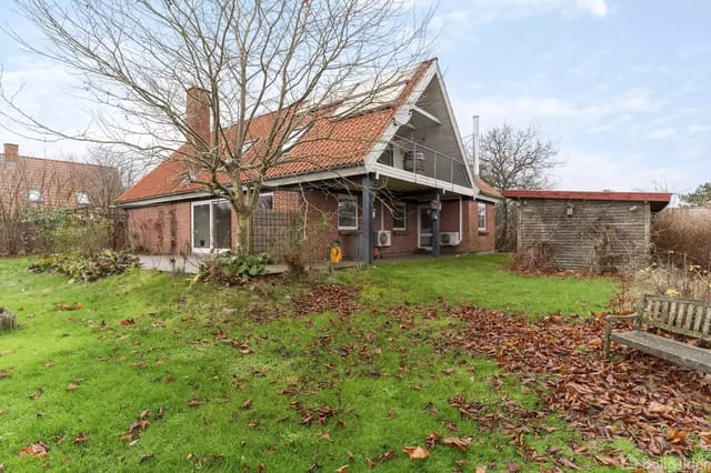 A brick house with a red tiled roof surrounded by a green garden with scattered autumn leaves. A wooden bench is on the right, and a shed is next to the house.