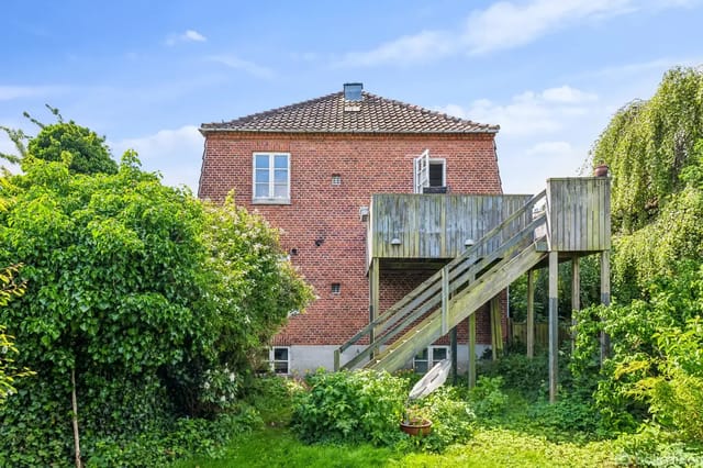 Red brick house surrounded by lush green plants, with a wooden staircase leading to an elevated terrace.