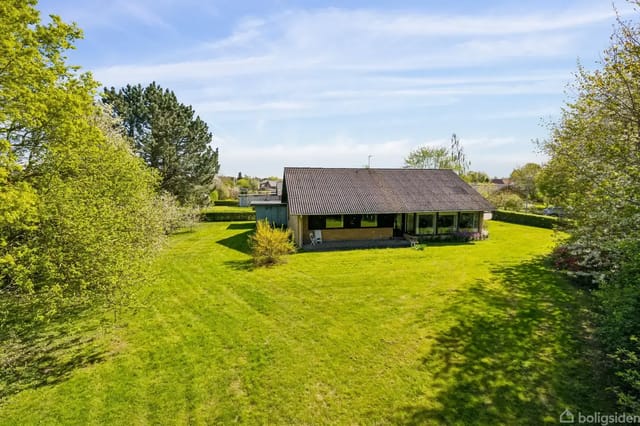 A house stands quietly in a large, green garden surrounded by trees and bushes. The lawn is well-kept under a blue sky with a few clouds.