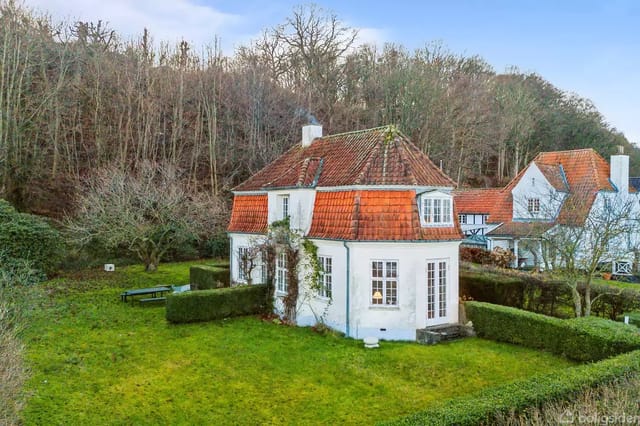 A charming white house with a red tile roof in a lush garden surrounded by bushes and trees, located near a forest.