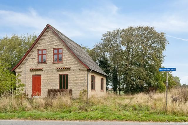 A small yellow brick house with red doors and windows stands quietly by a road, surrounded by tall grass and trees. Sign: 'Hagendrupvej'.