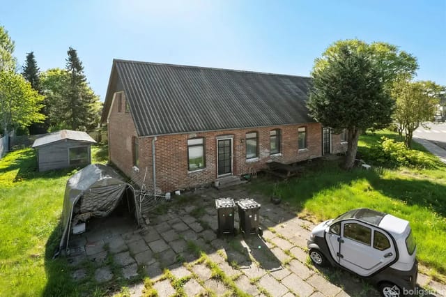 A brick building with a sloped roof and several windows stands next to a parked small electric car in a courtyard. The area is surrounded by grass and trees.