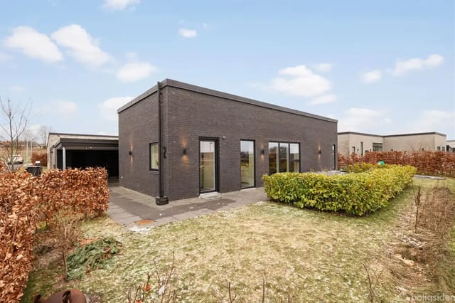 A modern, black brick building with large windows in a residential area. In front is a paved terrace and green bushes. The house has a carport on the left side.