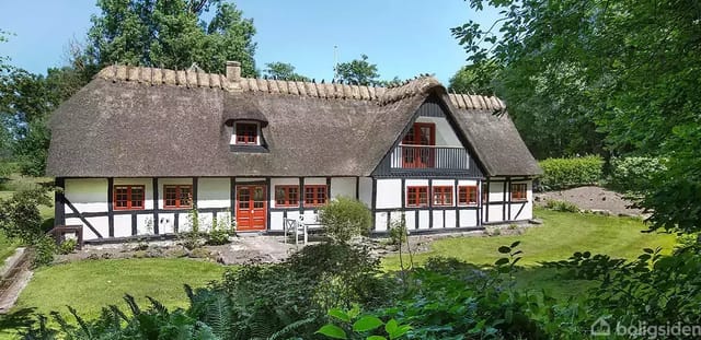 Traditional half-timbered house with thatched roof and red windows in a lush garden surrounded by trees and bushes.