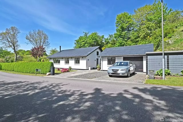 A white brick house with a black roof has a silver car parked in front on a gravel driveway. The house is surrounded by green trees and hedges under a clear blue sky.