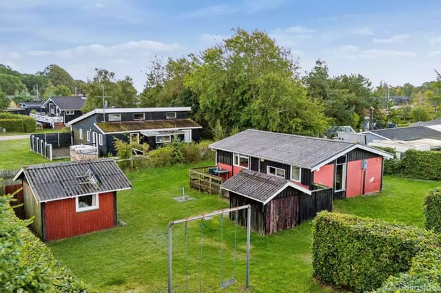 Red house with wooden cladding stands still in a green garden with a swing set; surrounded by several trees and other small houses in a residential area.