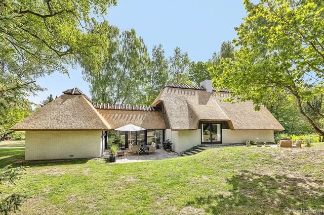 A thatched house stands in the middle of a lush garden. Sun loungers and a garden table with a parasol are placed on a terrace. The house is surrounded by trees and green areas.
