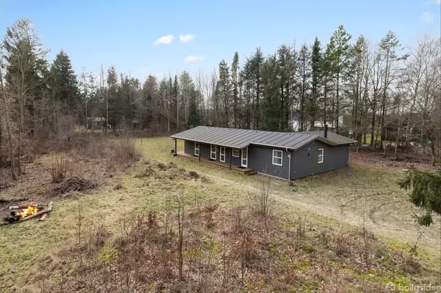 A gray house stands alone in a wooded field. Around the house are trees and bushes, and there is a bonfire with burning logs to the left.