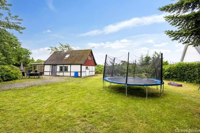 Trampoline on a lawn next to a traditional timber-framed house with red and white beams. Surrounding are bushes and trees, and a terrace with garden furniture.