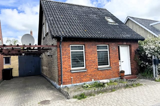 The house has a red brick facade with a black sloped roof and two front windows. Next to it is a carport. The entrance door is white. House number 25.