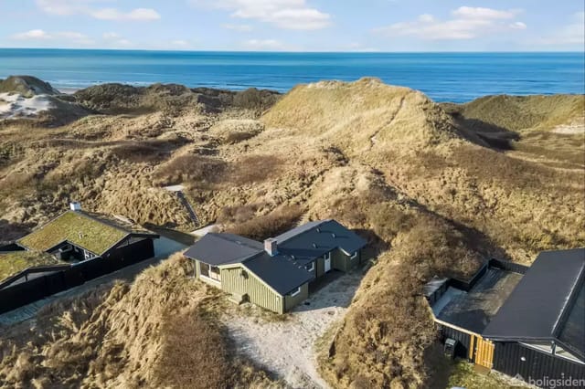 A modern house with a dark roof among grass-covered dunes near the coast. Other houses are surrounding. The sea stretches on the horizon under a blue sky with few clouds.