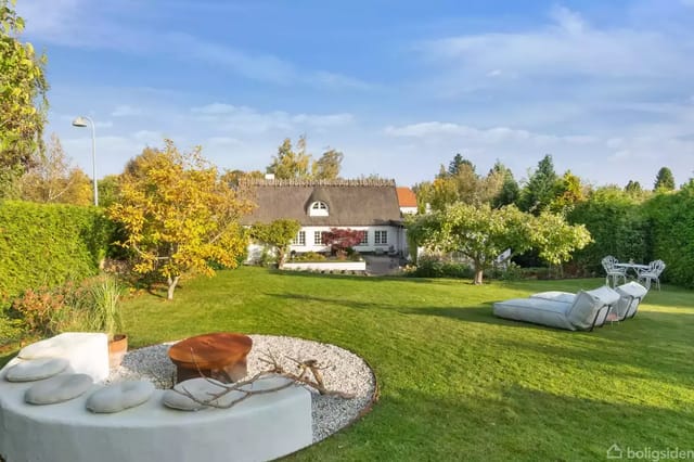 Thatched house in a green garden with lawn, bushes, and a seating area with concrete furniture.