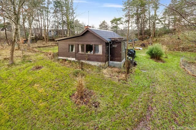 A small, brown wooden cabin stands quietly on a green, grassy plot surrounded by trees and bushes in a nature-rich area.