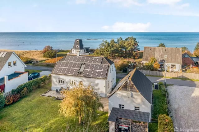 House with solar panels on the roof in a garden by the coast. The background shows a road, several houses, and the sea.