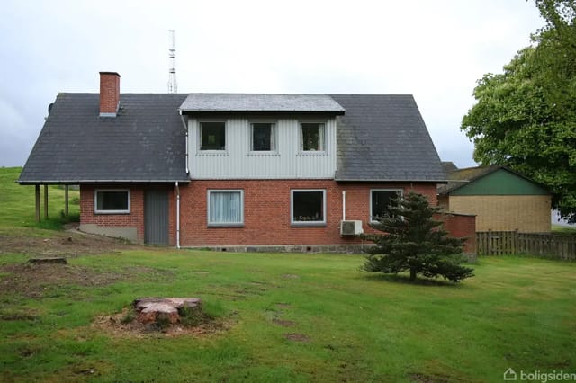 A red brick house with a black roof stands on a green lawn. There is a small fir tree in front of the house, with a tree stump to the left and a fence and tree to the right.