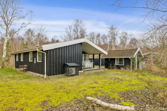 A black-painted summer house in a wooded garden with grass and dense trees around. A veranda with white furniture and a small shed in the background.