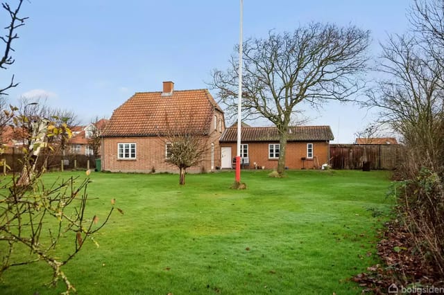 A red brick house with a tiled roof stands in a green, grassy garden with a large tree and a flagpole. Surrounded by fences and bushes, several buildings are seen in the background.